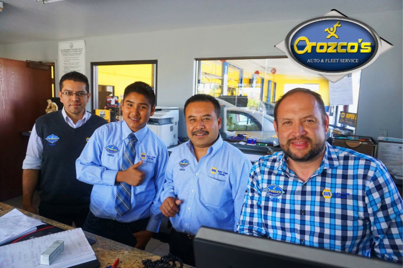 A teenage Andrew Orozco in an Orozco's Auto Service shirt and tie behind the service counter, with his father Servando and two colleagues