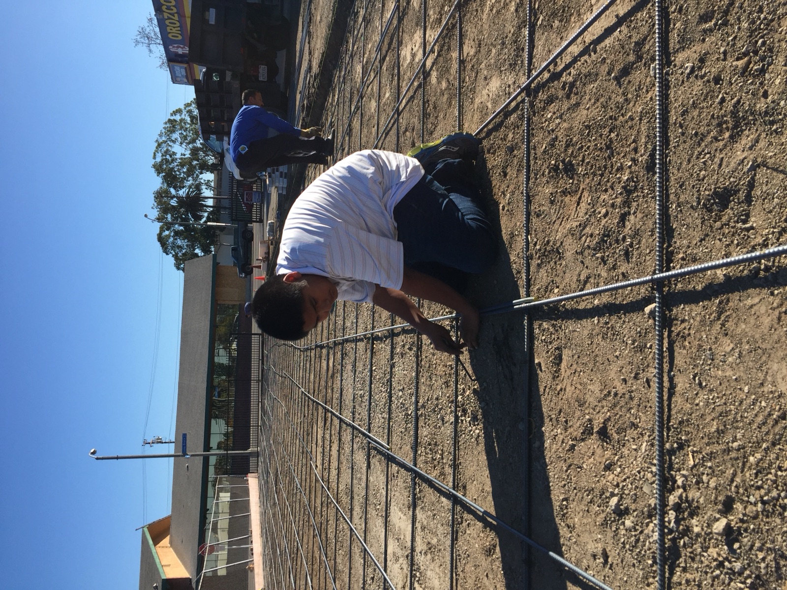 A young Andrew Orozco kneeling on a rebar grid, tying rebar while the parking lot at one of the family's shops is rebuilt, with an Orozco's truck behind him